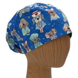 May include: Blue surgical scrub hat with a pattern of cartoon dogs wearing doctor and nurse costumes. The dogs are wearing stethoscopes, surgical masks, and hats. Some of the dogs have hearts and the word "LOVE" on their outfits.