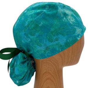 May include: A teal and green patterned scrub cap with a dark green ribbon tie. The cap is made of a soft fabric and is designed to be worn on the head. The cap is shown on a wooden mannequin head.