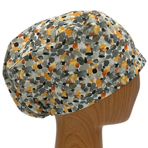 May include: A white scrub hat with a colorful pattern of gray, orange, and black pebbles. The hat is on a wooden mannequin head.