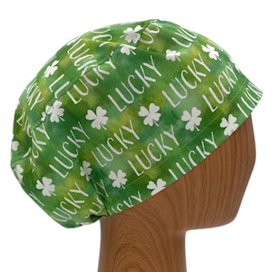 May include: Green fabric cap with white text and shamrock designs. The word "LUCKY" is repeated across the cap. The cap is on a wooden head form.
