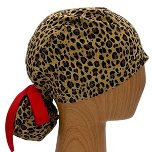 May include: A brown and gold leopard print surgical scrub hat with a red ponytail holder. The hat is on a wooden mannequin head.