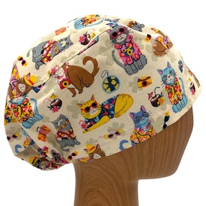 May include: A white scrub hat with a colorful pattern of cats wearing sunglasses and Hawaiian shirts. The cats are surrounded by palm trees, flowers, and other tropical imagery.