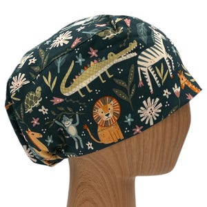 May include: A dark green scrub hat with a colorful jungle animal print. The print features a lion, zebra, crocodile, turtle, monkey, and giraffe. The hat is on a wooden mannequin head.