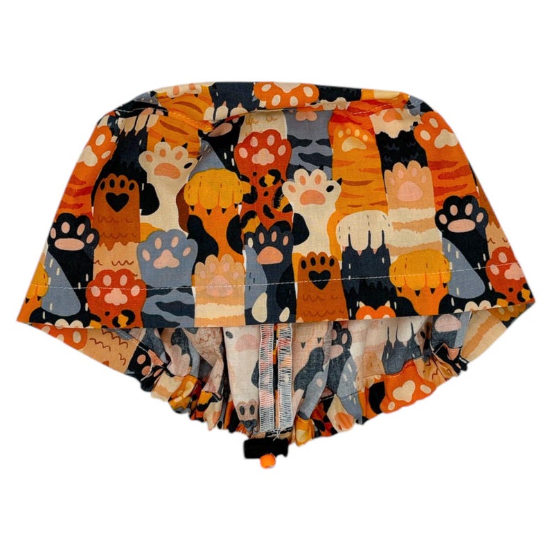 May include: A colorful surgical scrub hat with a paw print pattern in shades of orange, brown, gray, and black. The hat has a gathered elastic band at the bottom.