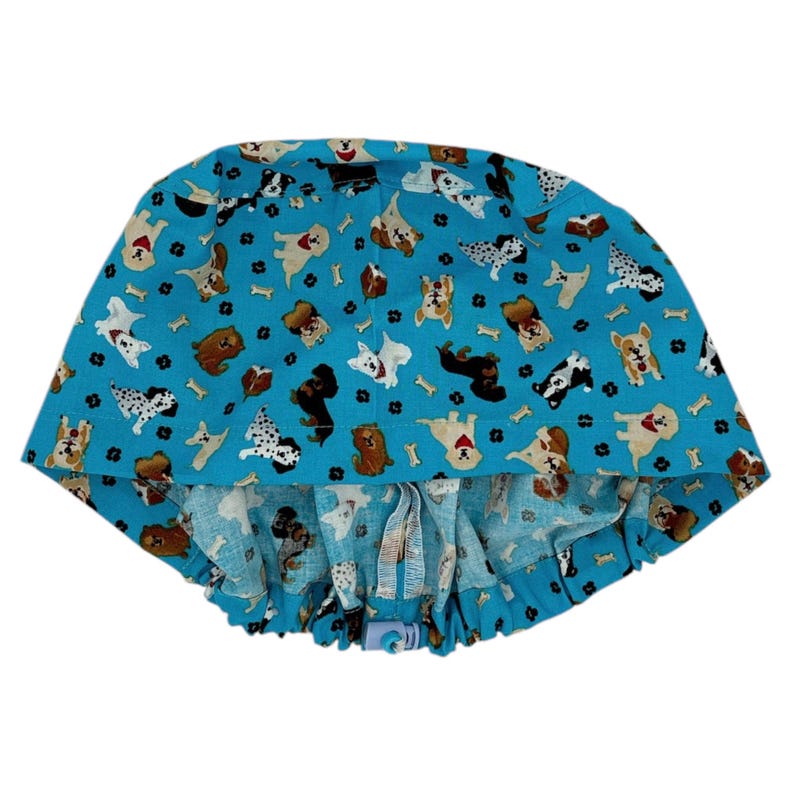 K&ouml;nnte beinhalten: Eine blaue OP-M&uuml;tze mit einem Muster aus Hunden und Knochen. Die M&uuml;tze hat einen Kordelzug und einen Knopfverschluss.