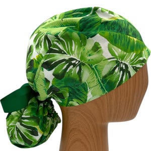 May include: Green and white surgical scrub hat with a tropical leaf print. The hat has a ponytail opening and a green ribbon tie.
