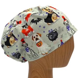 May include: A gray scrub hat with a Halloween-themed print featuring dogs dressed in costumes. The print includes dogs wearing a witch's hat, a vampire costume, a pirate costume, and a pumpkin costume. The hat has a star pattern and a black cauldron.