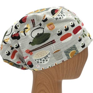 May include: A gray scrub hat with a sushi and Japanese food print. The hat features images of sushi rolls, chopsticks, teapots, and bowls of rice.