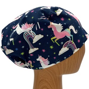 May include: A navy blue scrub hat with a whimsical unicorn print. The hat features pink, white, and rainbow colors on a dark blue background.