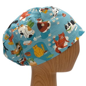 May include: A blue scrub hat with a pattern of cats and paw prints. The hat is on a wooden mannequin head.