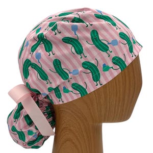 May include: Pink and white striped surgical scrub hat with a pattern of green smiling pickles holding blue balloons.