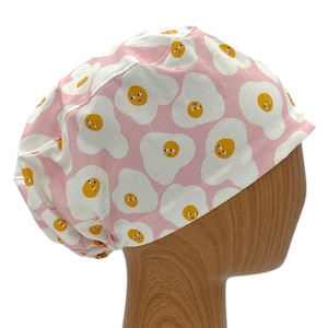May include: Pink scrub hat with a white and yellow print of smiling fried eggs.