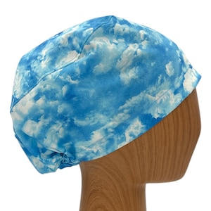 May include: A blue and white patterned scrub cap with a sky and cloud design. The cap is made of fabric and is designed to be worn on the head. The cap is shown on a wooden head form.