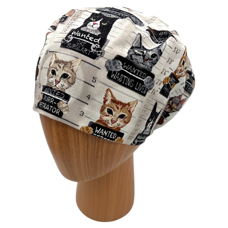 Puede incluir: Un gorro quir&uacute;rgico blanco con un patr&oacute;n repetido de fotos policiales de gatos con la palabra "WANTED" en letreros. Los gatos est&aacute;n representados en varios colores y poses, con textos como "CAT BURGLARY" y "WASTING LIVES".