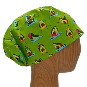 May include: Green scrub hat featuring a pattern of cartoon avocados performing yoga poses. The hat is displayed on a wooden mannequin head.