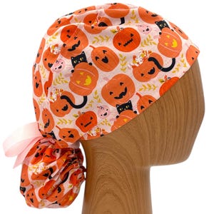 Könnte beinhalten: Eine OP-Haube im Halloween-Design mit orangefarbenen Kürbissen und schwarzen Katzen auf hellrosa Grund. Die Haube ist mit einem hellrosa Band gebunden.