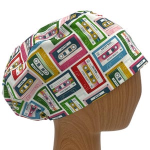 May include: A white surgical scrub cap with a colorful pattern of retro cassette tapes in various colors, including pink, yellow, green, and blue. The cap is designed to fit comfortably and securely.