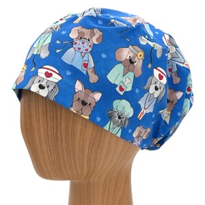 May include: Blue surgical scrub hat with a pattern of cartoon dogs wearing medical scrubs and hats. The dogs are wearing stethoscopes, bandages, and other medical accessories. The hat has a red heart pattern.