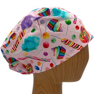 May include: Pink surgical scrub hat with a colorful candy print. The print features lollipops, candy canes, cotton candy, and other sweet treats.