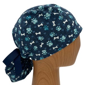 May include: A blue scrub hat with a pattern of paw prints and bones. The hat has a ponytail hole and a navy blue ribbon tie.