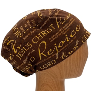 以下が含まれることがあります： 聖書の言葉が金色でプリントされた茶色のスクラブハット。"イエス・キリスト"、"喜びなさい"、"神を信頼しなさい"などの言葉が書かれています。