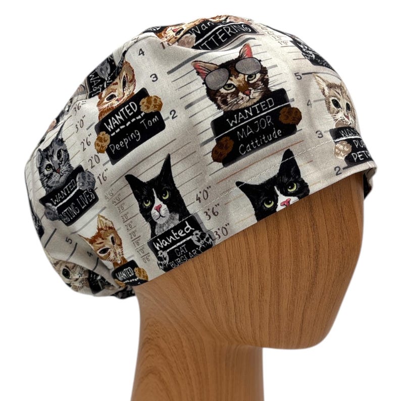 Puede incluir: Un gorro quir&uacute;rgico beige claro con un patr&oacute;n repetido de gatos de dibujos animados que sostienen letreros de "Se busca". Los letreros dicen "Peeping Tom", "Major Cattitude" y "Cat Burglar".