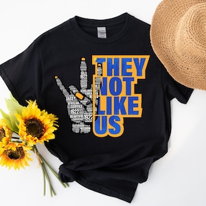 Peut inclure: T-shirt noir avec l'inscription "THEY NOT LIKE US" en bleu et jaune. Le signe de la paix est rempli de mots comme "SASSY" et "BOLD". Un chapeau de paille et des tournesols sont en arrière-plan.