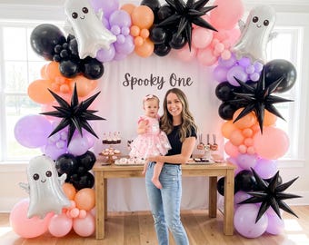May include: A balloon arch featuring black, orange, purple, and pink balloons, silver ghost-shaped foil balloons, and black starburst balloons. A white backdrop displays the text Spooky One. A wooden table holds various treats and desserts.