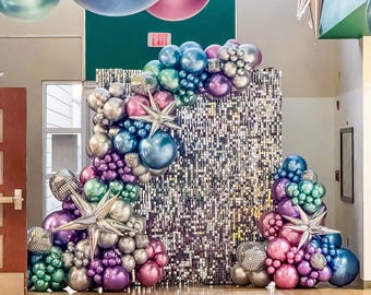 May include: A silver sequin backdrop with a balloon arch made of iridescent purple, pink, blue, and green balloons. The arch is decorated with silver stars.