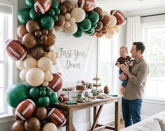 May include: A balloon arch in shades of dark green, brown, and cream, featuring football-shaped foil balloons. A white banner with the text First Year Down hangs above a wooden table displaying various treats and small football decorations.