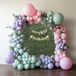 May include: A colourful balloon arch in pastel shades of pink, purple, and mint green, set against a green hedge backdrop. A gold banner reading "fairy 1st birthday" is suspended in the centre, creating a whimsical party scene.