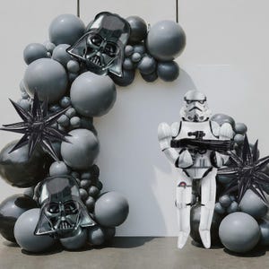 May include: A Star Wars-themed balloon arch with gray and black balloons, Darth Vader and star-shaped foil balloons. A Stormtrooper balloon stands in front of the arch, holding a blaster. Perfect for a Star Wars party.