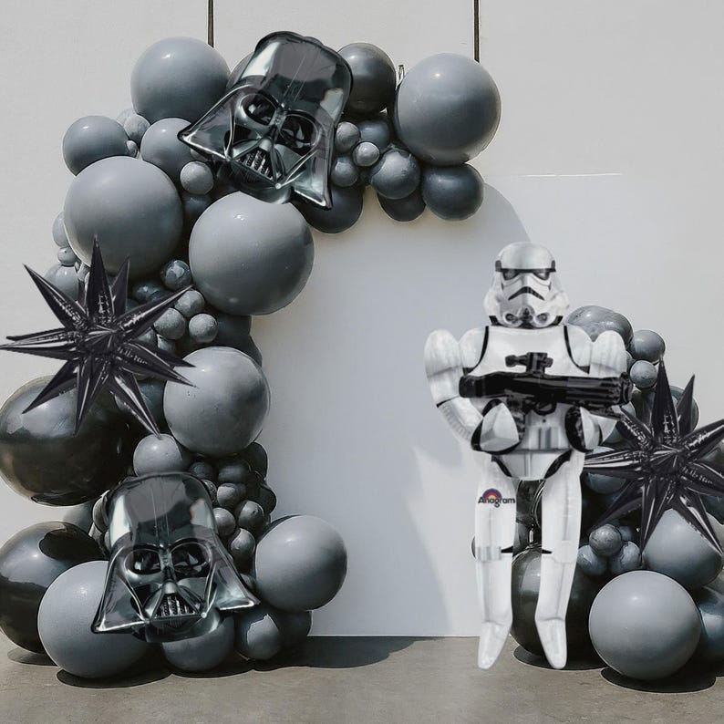 Może przedstawiać: Łuk balonowy o tematyce Gwiezdnych Wojen z szarymi i czarnymi balonami, Darth Vaderem i balonami foliowymi w kształcie gwiazd. Balon Stormtroopera stoi przed łukiem, trzymając blaster. Idealny na imprezę Gwiezdnych Wojen.