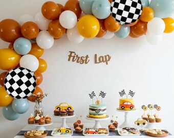 May include: A race car themed party dessert table with a balloon arch made of white, yellow, orange, and blue balloons. The arch has a checkered black and white balloon. The table has a cake with a checkered black and white design and a yellow race car on top. There are cupcakes with yellow race cars on top and cookies with yellow race cars on top. The table has a sign that says "First Lap".