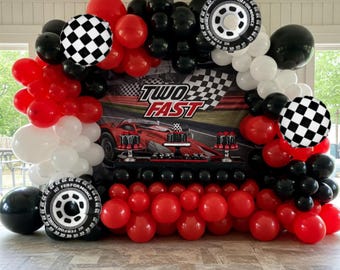 May include: A red, black, and white balloon arch with checkered and tire-shaped balloons. The arch is in front of a backdrop with a red race car and the words "Two Fast".