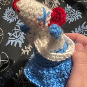 May include: A crocheted doll with a white body, red hat, and blue dress. The doll is wearing a blue and white cross on its chest.