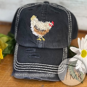 May include: A black baseball cap with a distressed finish and white contrast stitching. The front features an embroidered chicken with white, tan, and brown feathers, a red comb, and yellow feet. The text 2023ThreadedbyMaryKaye is visible.