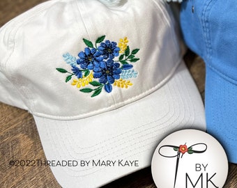 May include: White baseball cap with blue and yellow embroidered flowers. The cap has a curved brim and a white adjustable strap. The text "2022 THREADED BY MARY KAYE" is visible on the cap. A white circle with a red flower and the text "BY MK" is visible in the foreground.