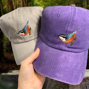May include: Two baseball caps, one tan and one purple, are displayed. Each cap features an embroidered bird design with blue, orange, and white details. The caps have a curved brim and an adjustable strap at the back.