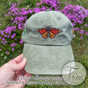 May include: An olive green baseball cap featuring an embroidered monarch butterfly on the front panel. The butterfly has orange, black, and white wings. The cap has a curved brim and stitched eyelets on the crown. The text 2026ThreadedbyMaryKaye is visible.