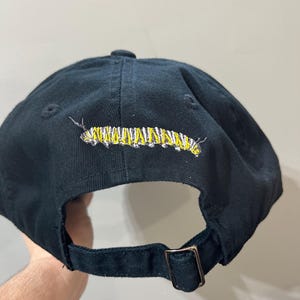 Peut inclure: Casquette de baseball noire avec une chenille jaune et blanche brodée à l'arrière.