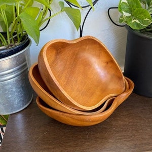 Pode incluir: Três tigelas de madeira, aninhadas, são feitas de madeira marrom claro. As tigelas têm formato de coração e têm um acabamento liso e polido.