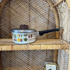 Może przedstawiać: Biały emaliowany garnek z czarną rączką. Garnek ma kolorowy wzór z kwiatami, owocami i przyborami kuchennymi. Garnek stoi na wiklinowej półce.