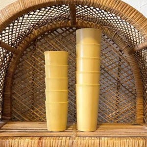 May include: Two stacks of yellow plastic tumblers are displayed on a wooden shelf. The tumblers are cylindrical with a slightly flared rim. The background features a woven wicker chair.