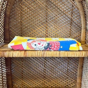May include: A folded, vintage pillowcase featuring a Raggedy Ann doll design. The pillowcase has a yellow background with a patchwork pattern and a smiling Raggedy Ann doll in the center. The doll has red hair, a white face, and a blue dress.