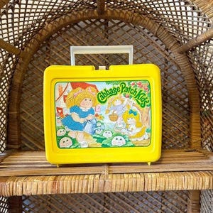 Op de afbeelding: Een gele Cabbage Patch Kids lunchbox met een witte handgreep. De voorkant heeft een kleurrijke illustratie van Cabbage Patch Kids-personages. De lunchbox is gemaakt van plastic en heeft een rechthoekige vorm.