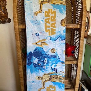 May include: A blue and white fabric panel featuring a Star Wars theme. The panel depicts characters from the Star Wars films, including Darth Vader, R2-D2, C-3PO, and Luke Skywalker. The panel also features the Star Wars logo in gold lettering.