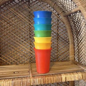 以下が含まれることがあります： 赤、黄、緑、ターコイズ、青の7色のプラスチックカップが積み重ねられています。カップは、編まれた籐の棚に積み重ねられており、籐の背景があります。