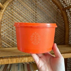 May include: An orange plastic container with a lid, held by a hand. The container has a decorative white floral design on the side. The container is cylindrical and appears to be vintage.