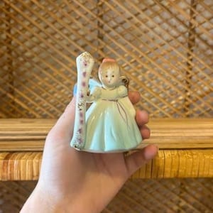 May include: A vintage porcelain figurine of a young girl angel in a pale green dress, holding a decorated staff. The figurine has a pink bow and gold accents. The angel has blue wings and is holding a staff with pink and gold floral designs.